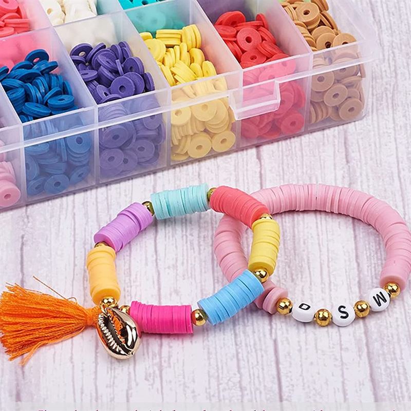 Two vibrant bracelets crafted with Fashiongirl’s KREA DIY Acrylic bead jewellery set lie beside its 24-compartment box of bright beads. One bracelet features an orange tassel and shell charm; the other spells BSW with white letter beads on a wooden surface.