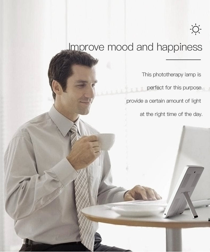 A man in a dress shirt sits at a table, holding a cup and using his laptop beside the Fashiongirl Light Therapy Lamp 10,000 lux LED. Text indicates that light therapy can help improve mood, happiness, and ease winter depression.