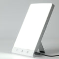 Fashiongirl Light Therapy Lamp 10 000 lux LED är en rektangulär lampa med en ljusvit skärm, utfällbart stöd, tre kontrollknappar och en strömsladd i botten – utformad för att simulera artificiellt dagsljus.