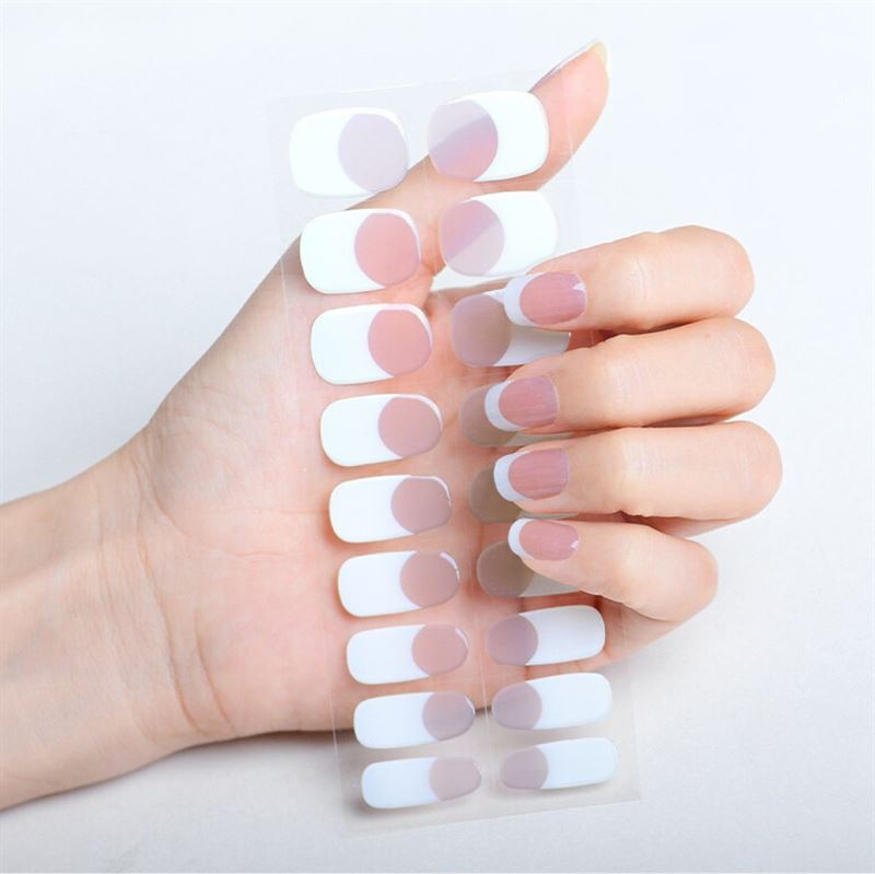 A hand with manicured nails holds UNIQ Semi Cured Gel Nail Stickers / Self-adhesive nail polish - Classic French (JK-331), featuring a chic white and light pink half-moon design that matches the nails perfectly.