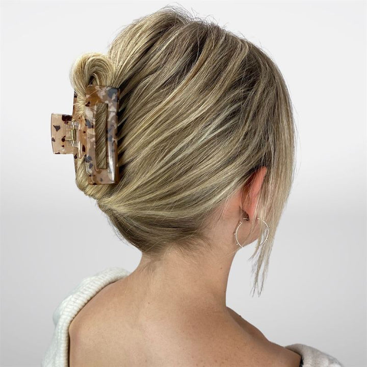 En kvinna med tjockt blont hår stylat i en låg fläta, fäst med SOHO Aria Hair Claw i Light Tortoise, bär silverörhängen och en vit topp, vänd bort från kameran mot en enfärgad bakgrund.