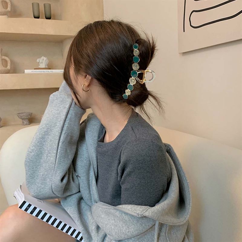 A woman with brown hair in a low bun, secured with the SOHO Becca Hair Claw - Ocean by SOHO, sits on a light couch wearing a grey sweater. She rests her hand on her face and looks away from the camera.