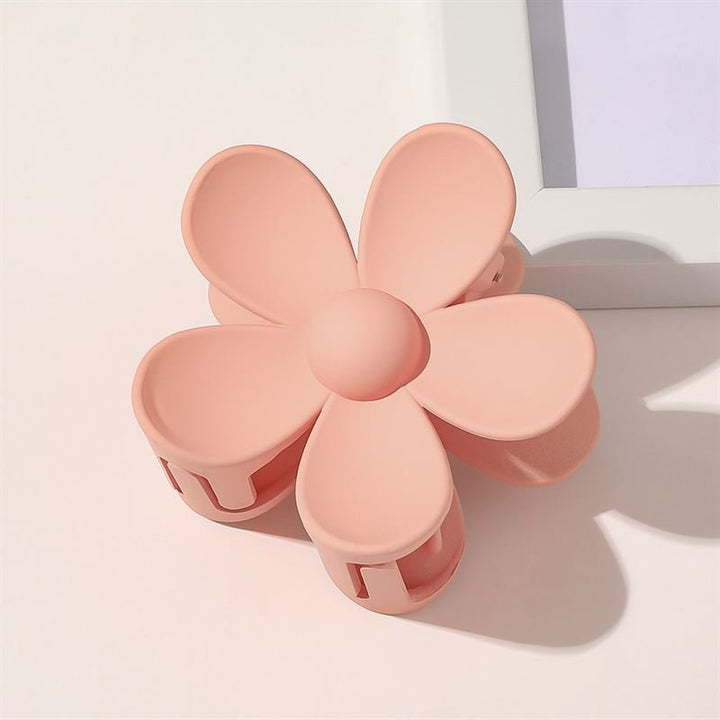 The SOHO Bloom Hair Claw - Peach by SOHO, shaped like a five-petaled pastel peach flower, sits on a white surface near the corner of a white picture frame.