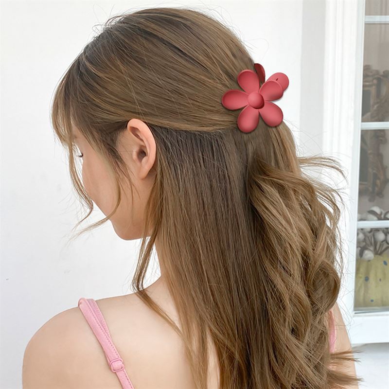 A woman with long, wavy brown hair is seen from behind wearing the SOHO Bloom Hair Claw - Pink Ombre by SOHO, paired with a light pink sleeveless top. A white wall and window are visible in the background.