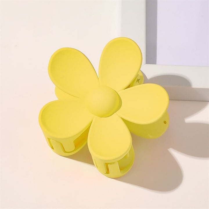 The SOHO Bloom Hair Claw - Yellow, a flower-shaped hair accessory by SOHO, rests on a light surface near the corner of a white picture frame.