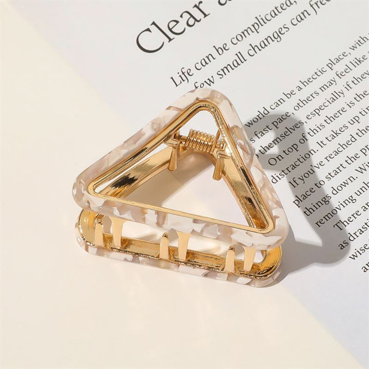 The SOHO Ea Hair Claw - Marble by SOHO, featuring a gold and white marbled design, rests on an open book or magazine as sunlight creates gentle shadows across the scene.