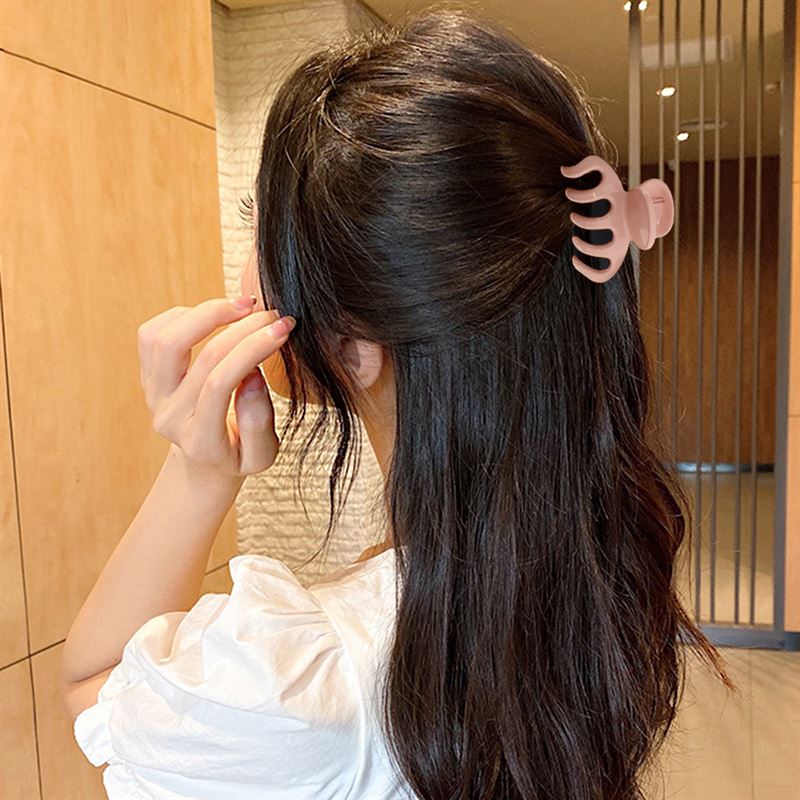 A woman with long dark hair, seen from behind, wears a white top and holds her left hand by her face. Her hair is partially clipped up with the SOHO Esta Hair Claw - White by SOHO. The background features wooden and white textured walls.