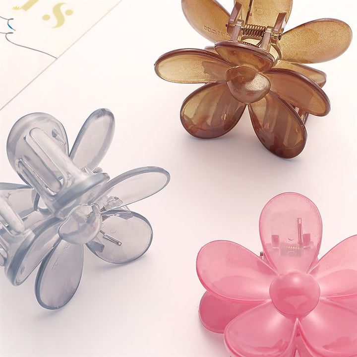 The SOHO Fiore Hair Claw - Charm Pink, from SOHO, features a petal-inspired design and is shown with translucent brown and gray versions, arranged on a light surface near the edge of white paper.