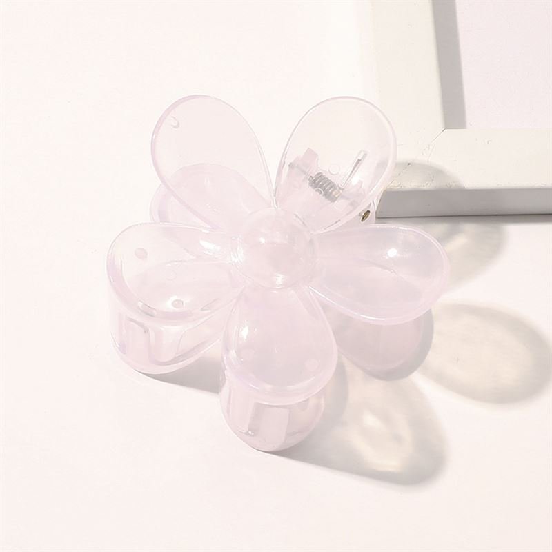 The SOHO Fiore Hair Claw in Pale Purple, a translucent six-petal flower design by SOHO, rests on a white surface beside the corner of a white picture frame.