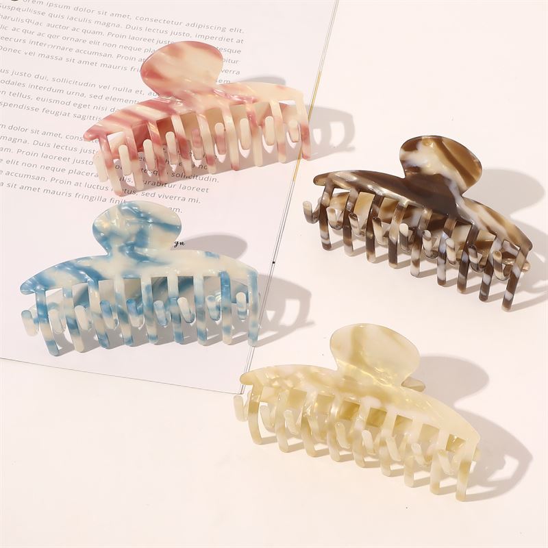 The SOHO Gaja Hair Claw in Beige Marble, along with pink, brown, and blue marbled accessories, are artfully arranged on a flat surface near an open magazine as sunlight creates soft shadows across this elegant SOHO collection.