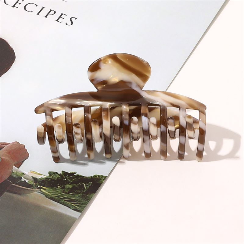 SOHO Gaja Hair Claw i brun marmor från SOHO ligger på en öppen tidning och kastar en stilfull skugga – ett elegant marmorerat tillbehör som är perfekt för att lyfta din vardagslook.