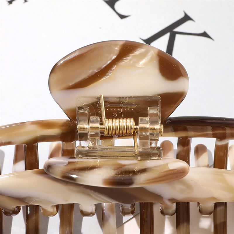 En närbild av SOHO Gaja Hair Claw - Brown Marble från SOHO, som framhäver dess fjädermekanism och böjda tänder mot en suddig bakgrund med delvis svart text.