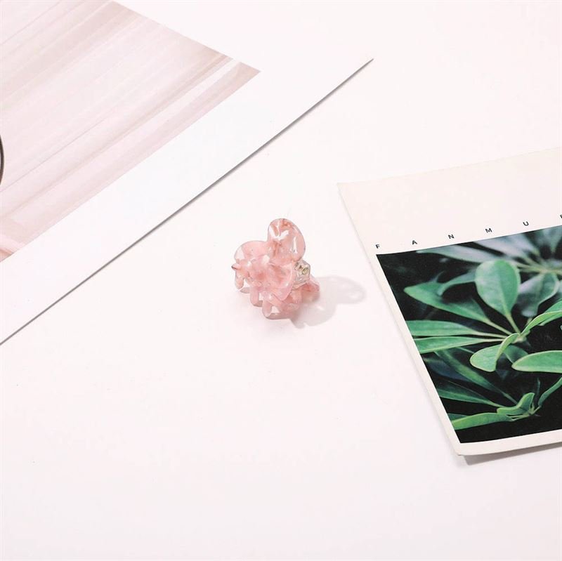 The SOHO Hara Mini Hair Claw - Blush, a pink translucent clip from SOHO, rests on a white surface between two papers—one with green leaves and the other with abstract pastel lines.