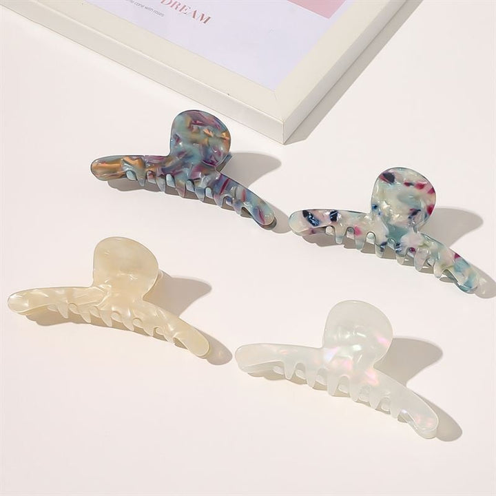 Four pastel SOHO hair claws, including the SOHO Haylen Hair Claw in white with marbled and pearlescent designs, are arranged on a white surface by a picture frame—an elegant and eye-catching accessory collection.