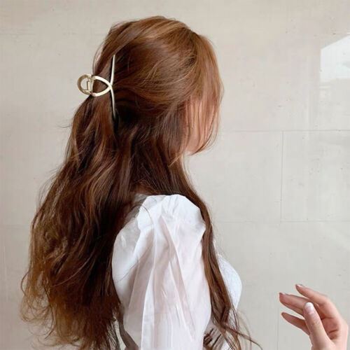 A woman with long, wavy brown hair, wearing a white top, secures her hairstyle with the SOHO Iva Metal Hair Claw in Silver by SOHO. She faces sideways as a hand holding a nail extension appears in the foreground against a pale wall.