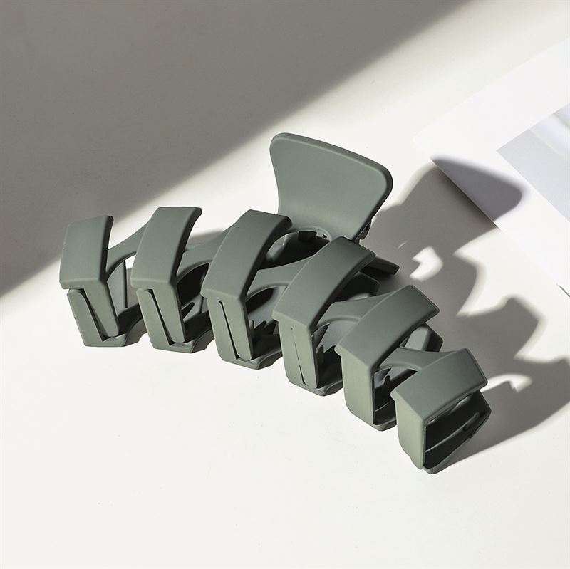 The SOHO Just Hair Claw - Green by SOHO, a large matte olive hair clip with wide rectangular teeth, sits on a white surface near partially shadowed paper showing a gray section.