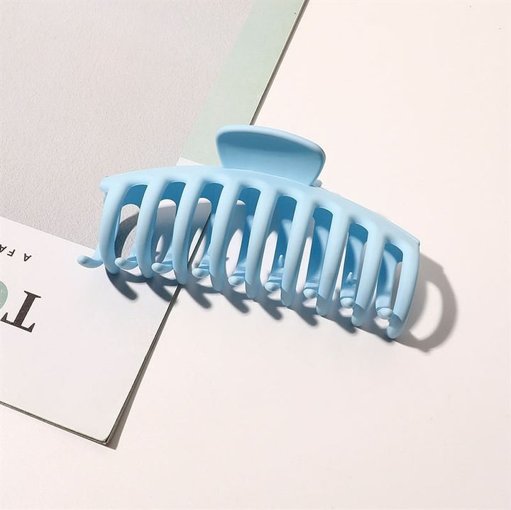 The SOHO Kana Hair Claw in light blue, by SOHO, features a strong grip and rests on a white surface near printed paper, casting a soft shadow. Suitable for all hair types.