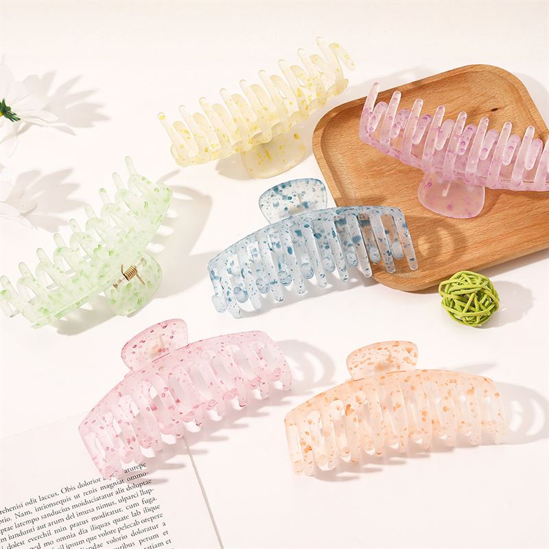 The SOHO Kana Splash Hair Claw in Blue by SOHO is a large, translucent clip with a strong grip, perfect for all hair types. Styled on a white surface with pastel clips, it adds a chic touch to any look. Magazine and decor not included.