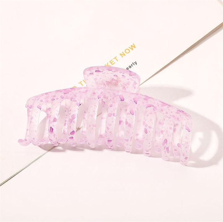 The SOHO Kana Splash Hair Claw - Purple by SOHO features a translucent pink design with purple speckles, shown resting on a white surface with printed text underneath. This clip provides a strong grip and suits all hair types.
