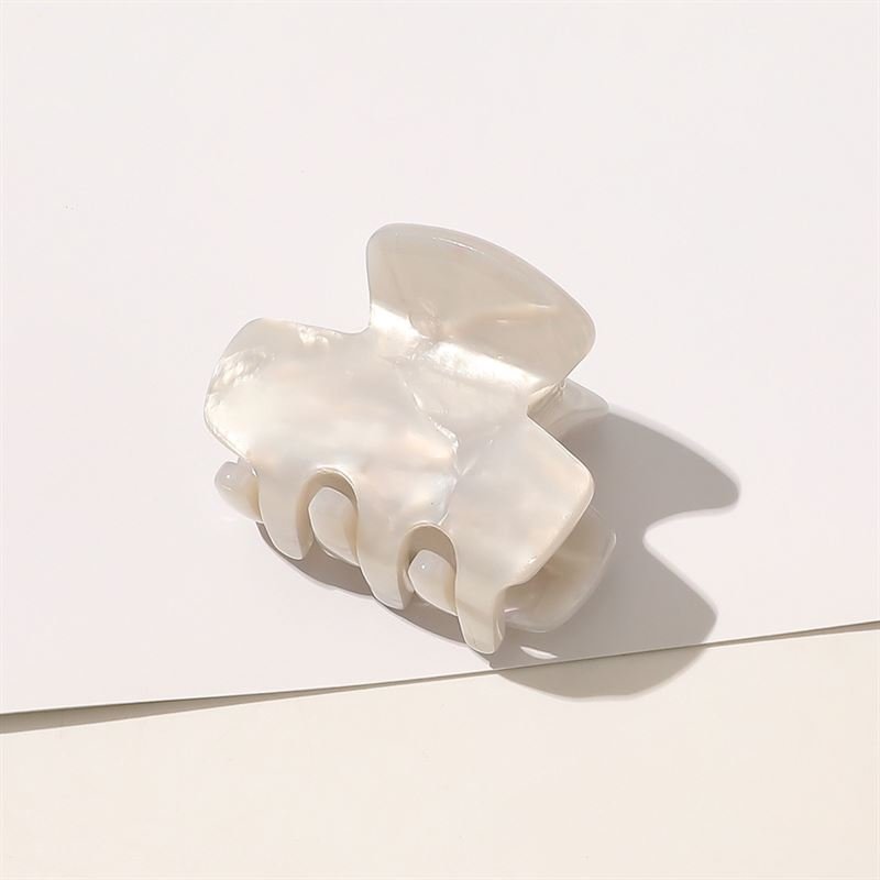 The SOHO Loa Mini Hair Claw in White Marble features curved prongs and a pearlescent shine, resting on a white surface with a soft shadow.