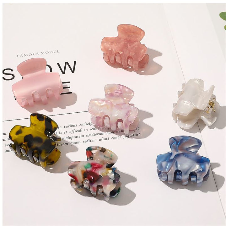 Seven colorful SOHO hair claws, including the SOHO Loa Mini Hair Claw in White Marble, are artfully arranged in different shapes and patterns on an open magazine page under bright, even lighting.