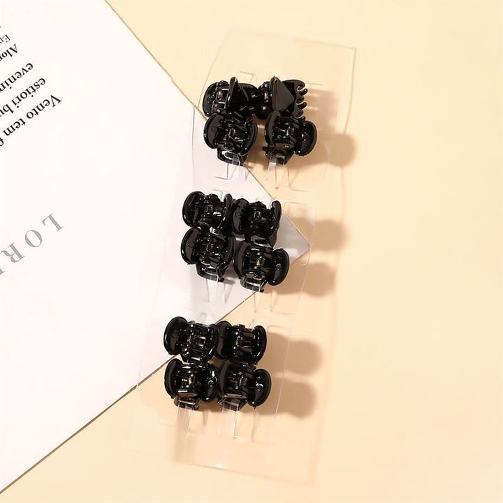 A clear plastic package of eight SOHO Lucca Mini Hair Claws - Black by SOHO rests on a light beige surface beside a partially visible sheet of paper with black printed text.