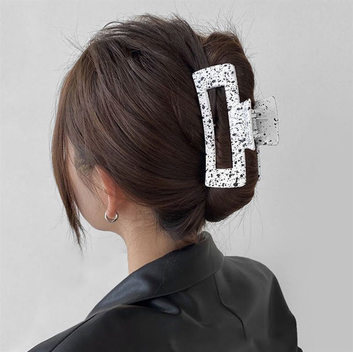 A woman with brown hair styled in a twist, secured with the SOHO Luva Hair Claw - White by SOHO. She is wearing a black jacket and small hoop earrings.