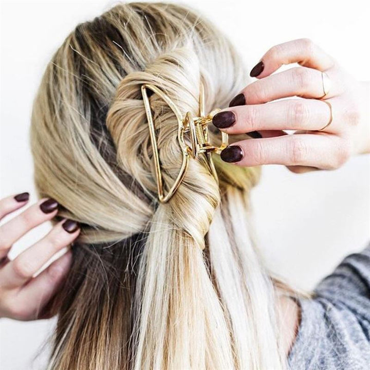 A person with blonde hair styles a twisted half-up look, securing it with the SOHO Lyra Large Metal Hair Claw in Gold by SOHO. They have dark-painted nails, a gray top, and wear a thin ring on one finger.