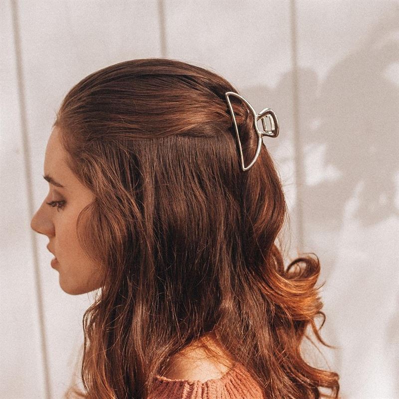 A woman with wavy brown hair styled half-up and secured with the SOHO Lyra Large Metal Hair Claw in gold, wearing a pink knit top, is shown in profile against a white paneled background.