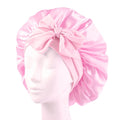 SOHO Makena Satin Bonnet - Pink från Fashiongirl, som visas på en mannekänghuvud, har en glänsande rosa satinfinish och en stor rosett framtill – perfekt för att skydda håret och förhindra frissighet.