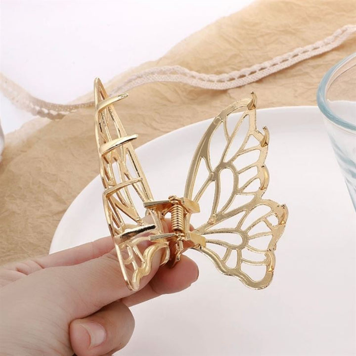 A hand holds the SOHO Metal Butterfly Hair Claw - Silver near a glass and a white plate on a light surface, with beige fabric and lace in the background. This SOHO accessory offers elegance and a secure grip for your hair.