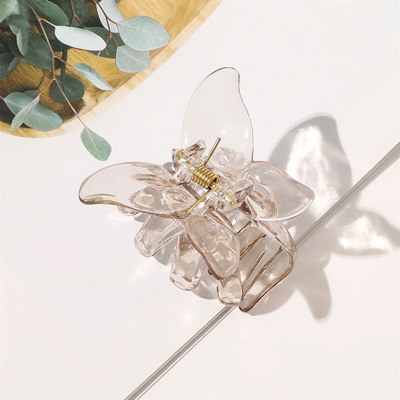 The SOHO Naja Hair Claw - Espresso by SOHO, featuring a translucent brown finish and gold spring, rests on a white surface with green eucalyptus leaves in the background, complementing this elegant hair accessory.