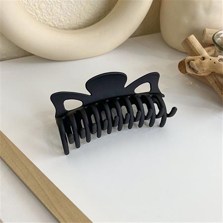 The SOHO Zara Hair Claw - Black by SOHO, featuring a matte finish and curved teeth, rests on a white surface with beige decor and wooden accents in the background.