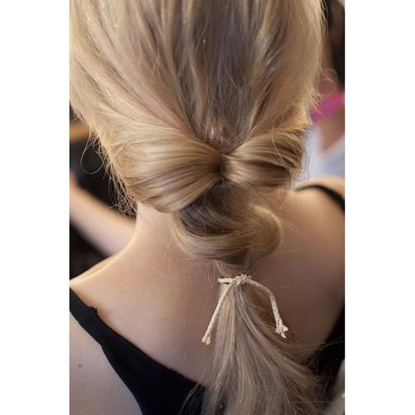 A woman with long, blonde hair styled in a creative bow-shaped low ponytail using the Fashiongirl Topsy Tail Hair Styling Tool 2-piece set. She wears a black thin-strap top, showcasing easy, stylish updos perfect for this versatile hair tool.