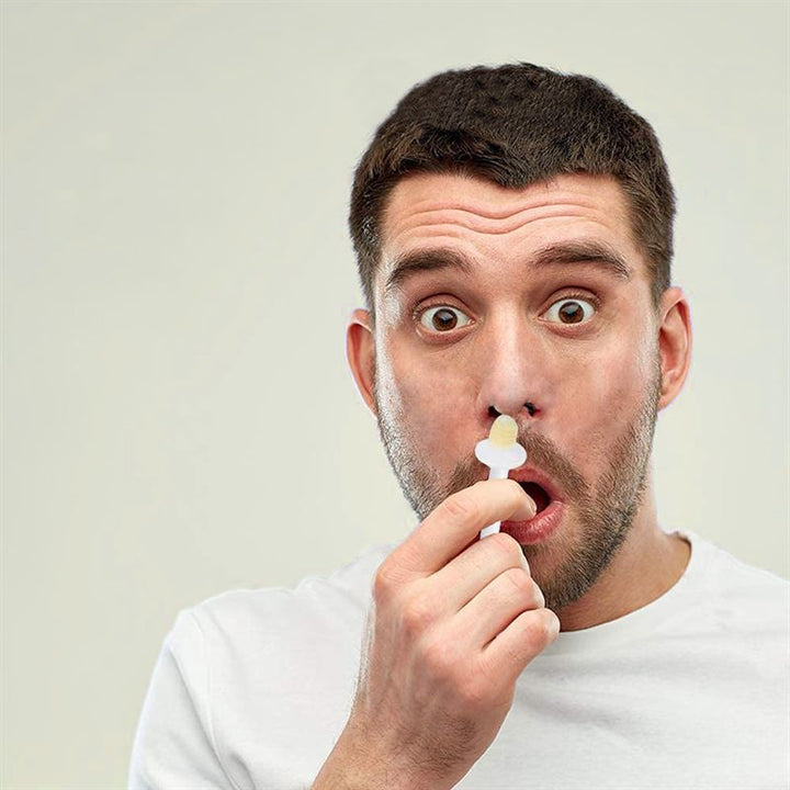 A surprised man in a white shirt holds the UNIQ Nose Wax Kit by UNIQ near his nose, seemingly about to use it for removing nose hair. The background is plain and light-colored.