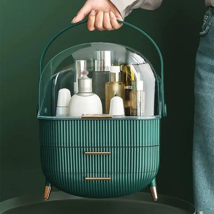 A person holds the UNIQ Retro Makeup Box Organiser in Dark Mint by UNIQ, featuring gold legs, two drawers, and a clear dome lid that reveals various skincare and cosmetic bottles.
