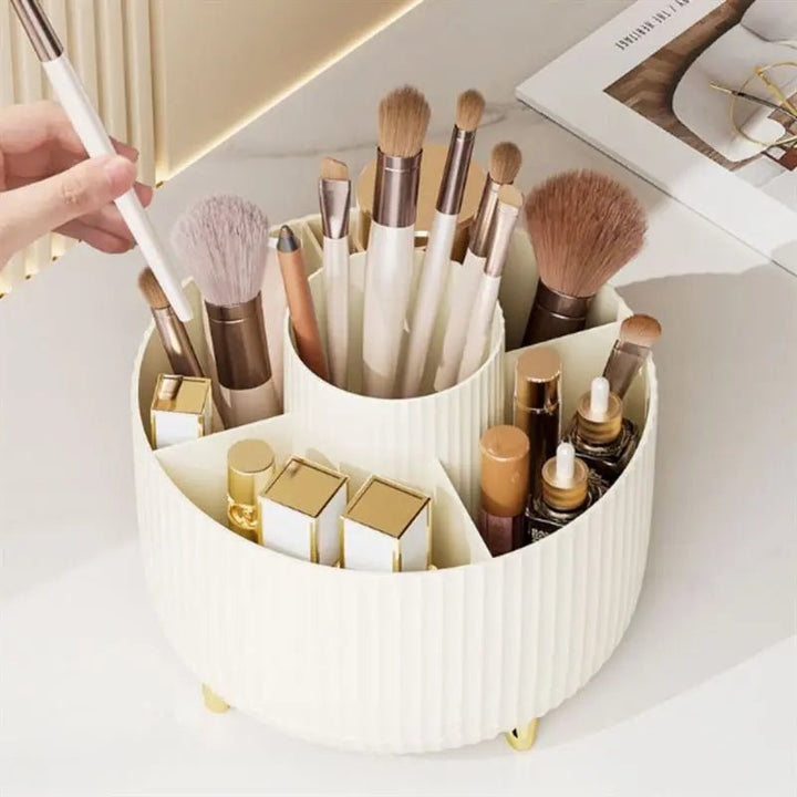 The UNIQ Rotating Makeup Organiser in Cream White by UNIQ sits on a vanity, holding brushes, lipsticks, and beauty products. A hand selects a makeup brush while a magazine and mirror are in the background.